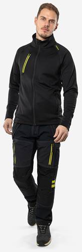 Eine Person, die eine schwarze Jacke und schwarze Hosen mit gelben Reißverschlüssen trägt, steht vor einem weißen Hintergrund.