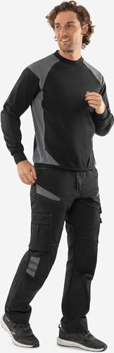 Eine Person in einem schwarzen und grauen Langarmshirt und schwarzer Hose ist von der Taille aufwärts zu sehen, wobei die rechte Hand sichtbar ist.