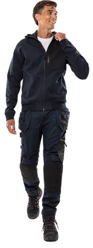 Eine Person trägt eine dunkle Jacke und Cargohose und steht mit leicht ausgestreckter linker Hand.