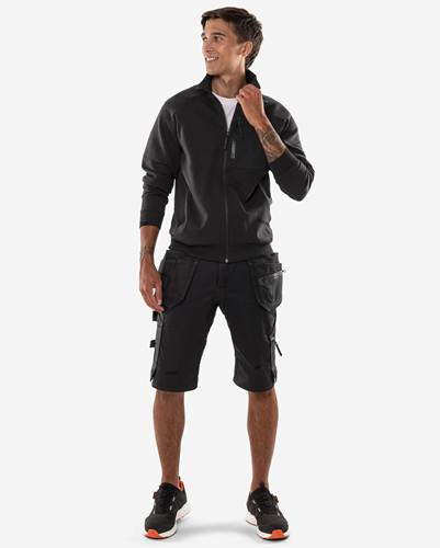 Eine Person steht und trägt eine schwarze Jacke und schwarze Cargo-Shorts, mit den Händen in den Taschen und blickt zur Seite.