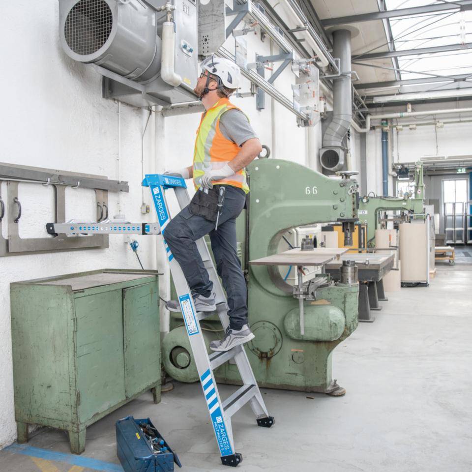 Ein Arbeiter in einer Sicherheitsweste und einem Schutzhelm steht auf einer Klappleiter und inspiziert Maschinen in einer Industriewerkstatt.