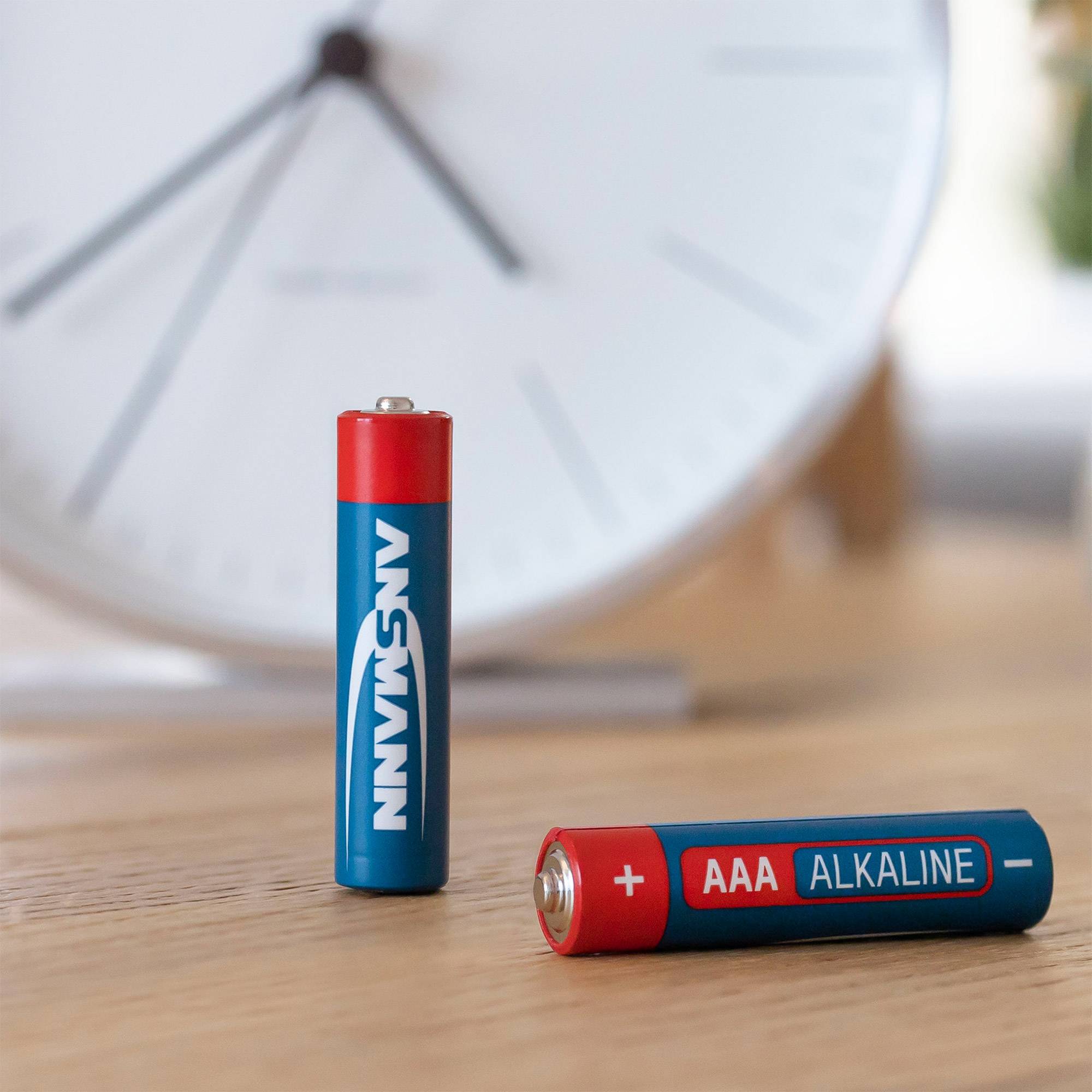 Zwei AAA-Alkalibatterien auf einer Holzoberfläche, eine aufrecht und eine liegend, mit einer unscharfen Uhr im Hintergrund.