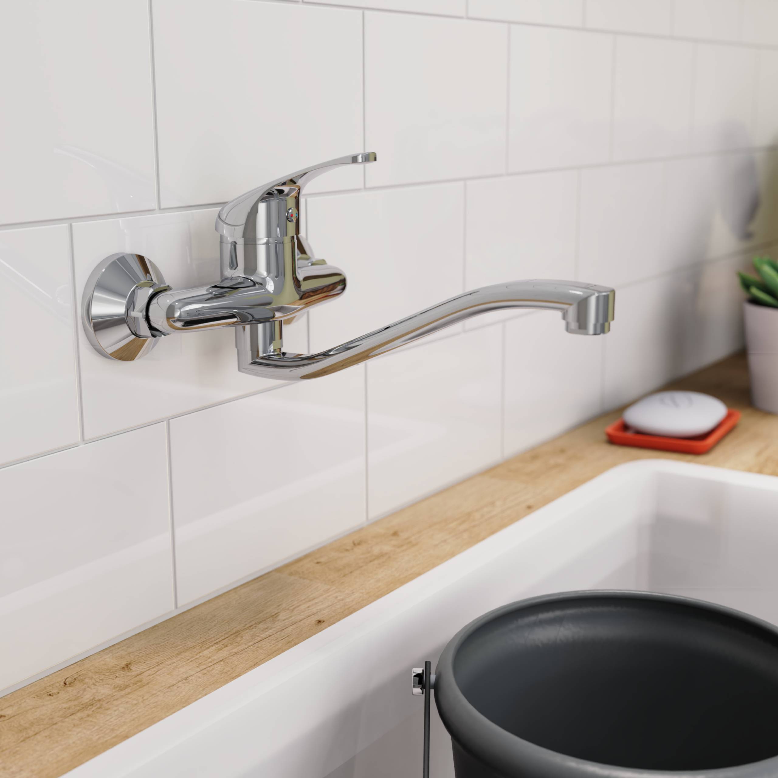 Ein moderner, verchromter Wasserhahn ragt über ein weißes Waschbecken mit einer Holzarbeitsplatte. Ein kleines rotes Seifenschälchen ist im Hintergrund zu sehen.