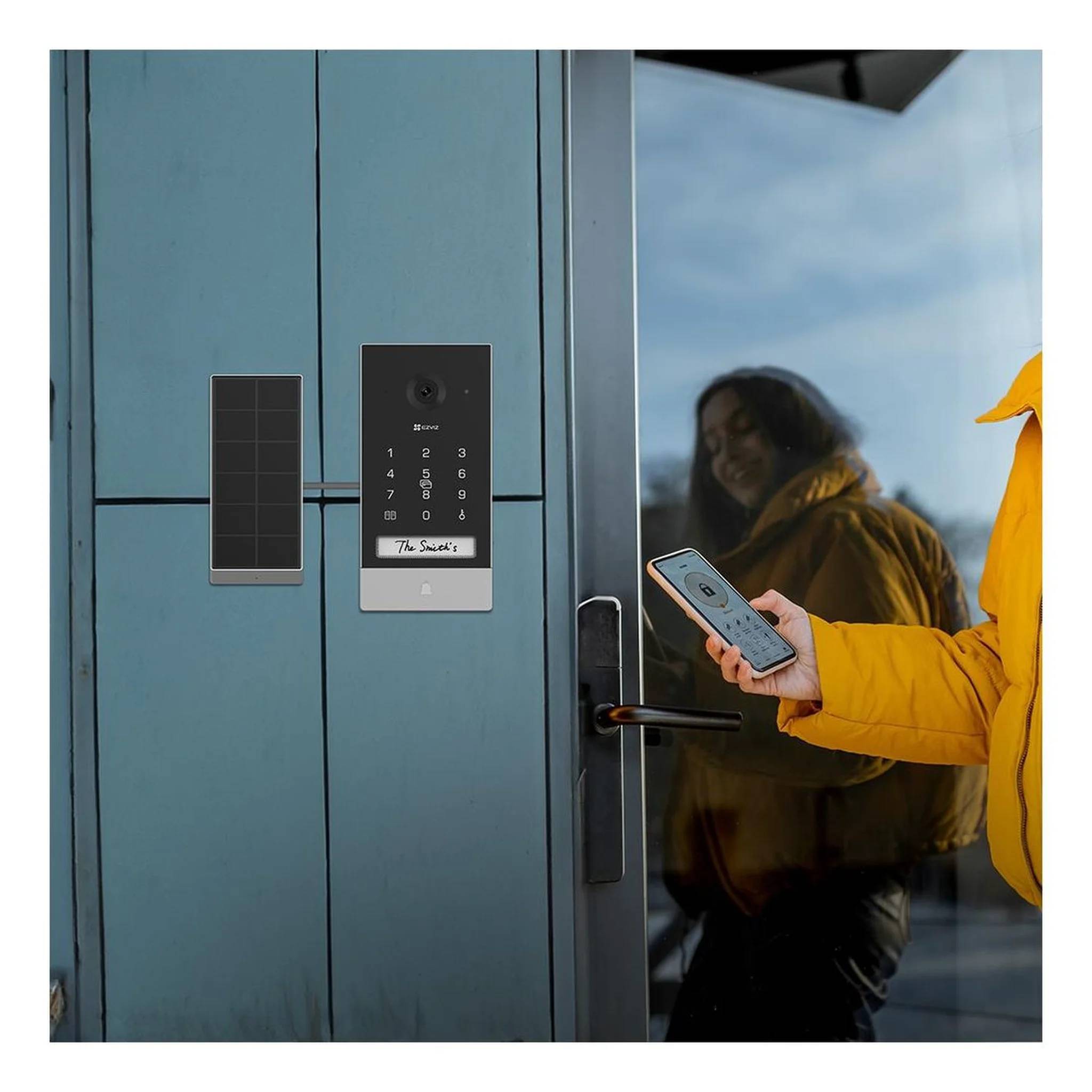 Eine Person in einer gelben Jacke benutzt ein Smartphone, um ein intelligentes Türöffnungssystem mit Tastatur und Kartenleser an einer blauen Tür freizuschalten.