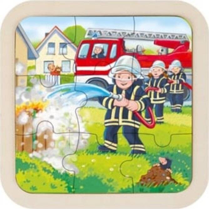 Feuerwehrmänner in Uniform sprühen Wasser aus einem Schlauch auf ein kleines Feuer außerhalb eines Hauses, mit einem Feuerwehrwagen und weiteren Feuerwehrmännern im Hintergrund.