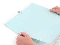 Hände, die ein hellblaues Blatt Papier auf eine Schneidematte oder Arbeitsfläche legen.