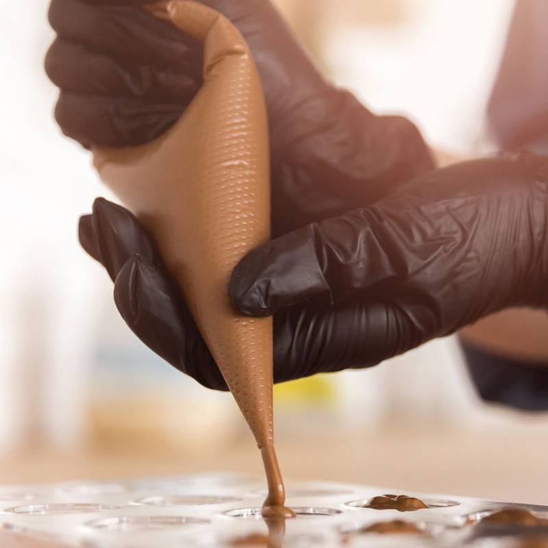 Eine Hand in einem schwarzen Handschuh spritzt Schokolade in kleine Förmchen und legt dabei besonderen Wert auf Präzision in einer Konditorei oder Backumgebung.