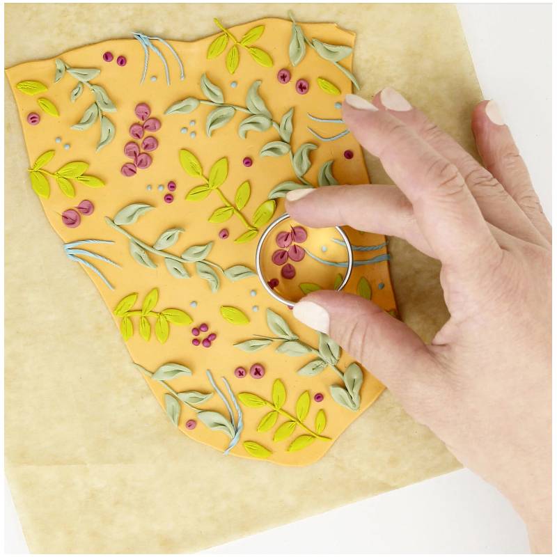 Eine Hand drückt einen runden Ausstecher in ein Blatt orangefarbenen Tons, das mit bunten Blumenmuster verziert ist und bereitet sich darauf, eine kreisförmige Form auszuschneiden.