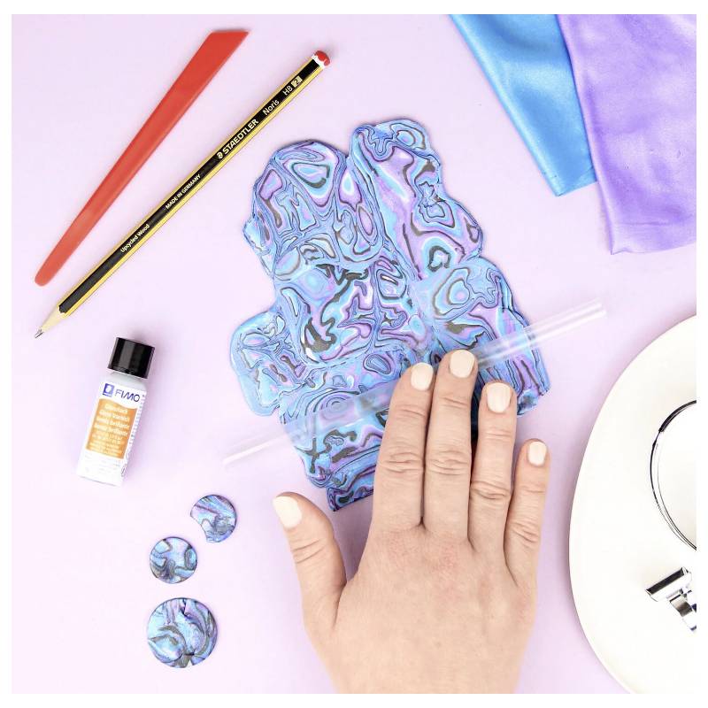 Eine Hand, die einen blauen marmorierten Tonbogen mit einer Rolle auf einer Lavendeloberfläche glättet, umgeben von Bastelmaterialien, einem Bleistift und Stoff.