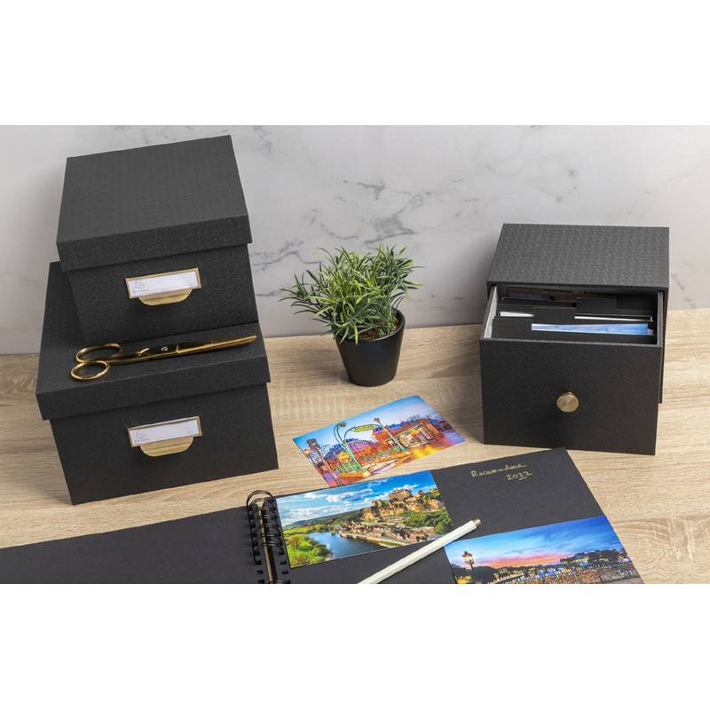 Ein Schreibtisch mit schwarzen Aufbewahrungsboxen, einer kleinen Topfpflanze, einem geöffneten Scrapbook mit Fotos, Postkarten und goldener Schere, was Organisation und Kreativität andeutet.