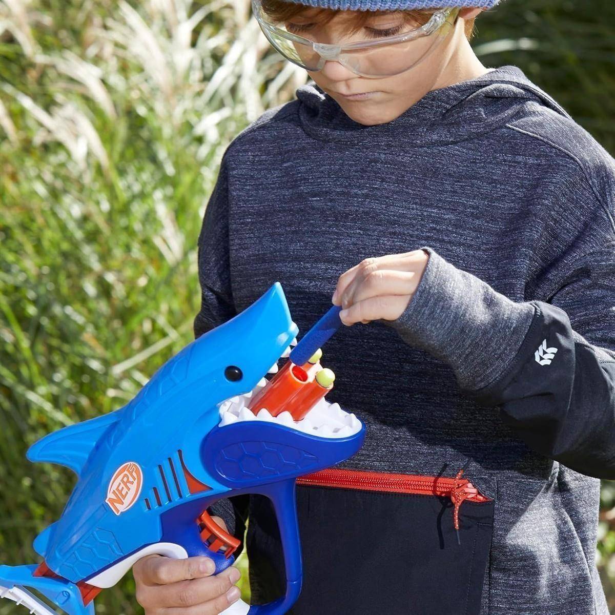 Kind mit Schutzbrille spielt draußen mit einer blauen Spielzeughai-Blaster-Pistole und lädt Pfeile in das offene Haimaul.