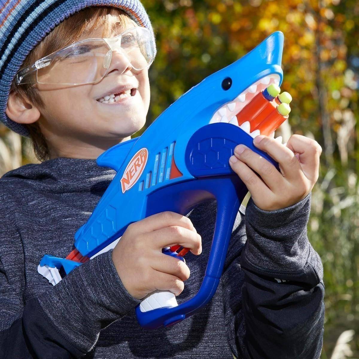 Kind mit Schutzbrille und gestreifter Mütze hält ein blaues haiförmiges Nerf-Spielzeuggewehr im Freien.