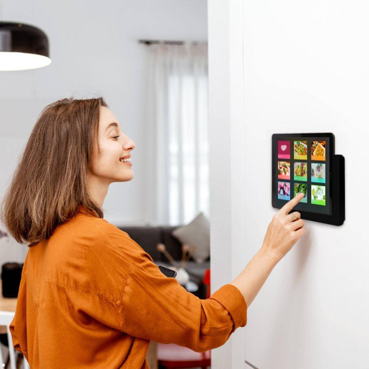 Eine Person in einem orangefarbenen Shirt interagiert mit einem an der Wand montierten Tablet, das bunte App-Symbole zeigt, in einem hellen, modernen Wohnumfeld.