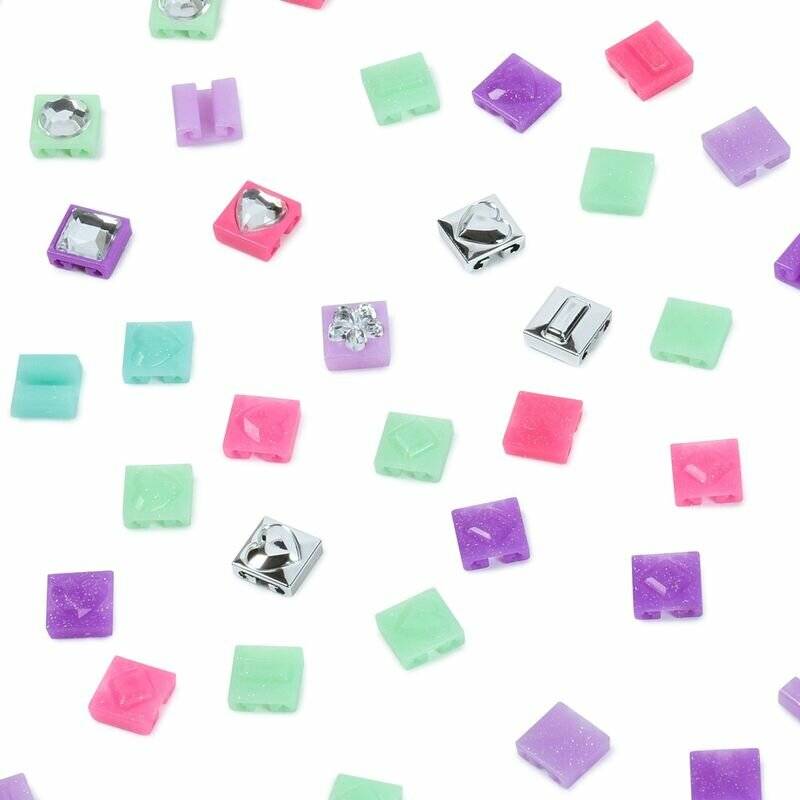 Assortierte kleine Perlen oder Anhänger aus Pastelltönen in Quadratform, verstreut auf weißem Hintergrund, in Mintgrün, Violett, Rosa und silberner Metallicoberfläche.