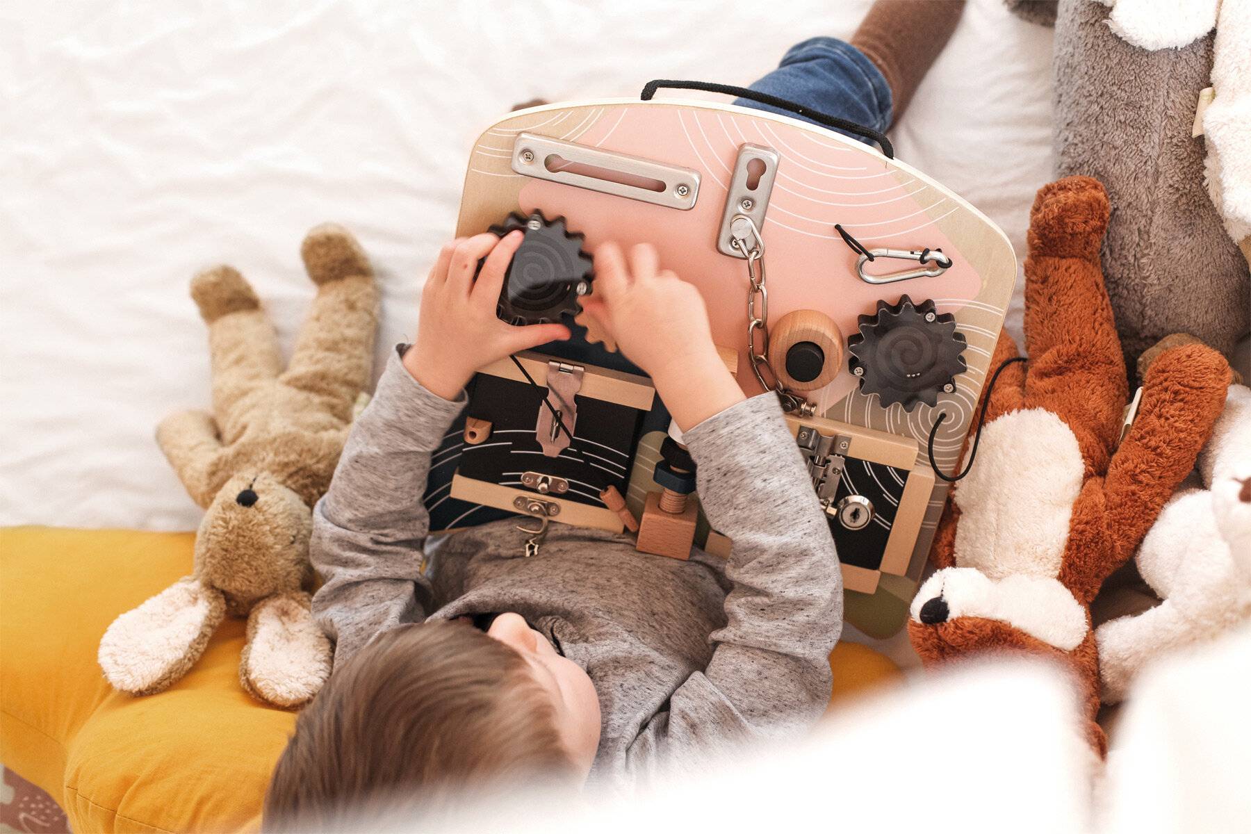 Kind auf einem Bett liegend, spielend mit einem hölzernen Beschäftigungsspielzeug, umgeben von Plüschtieren.