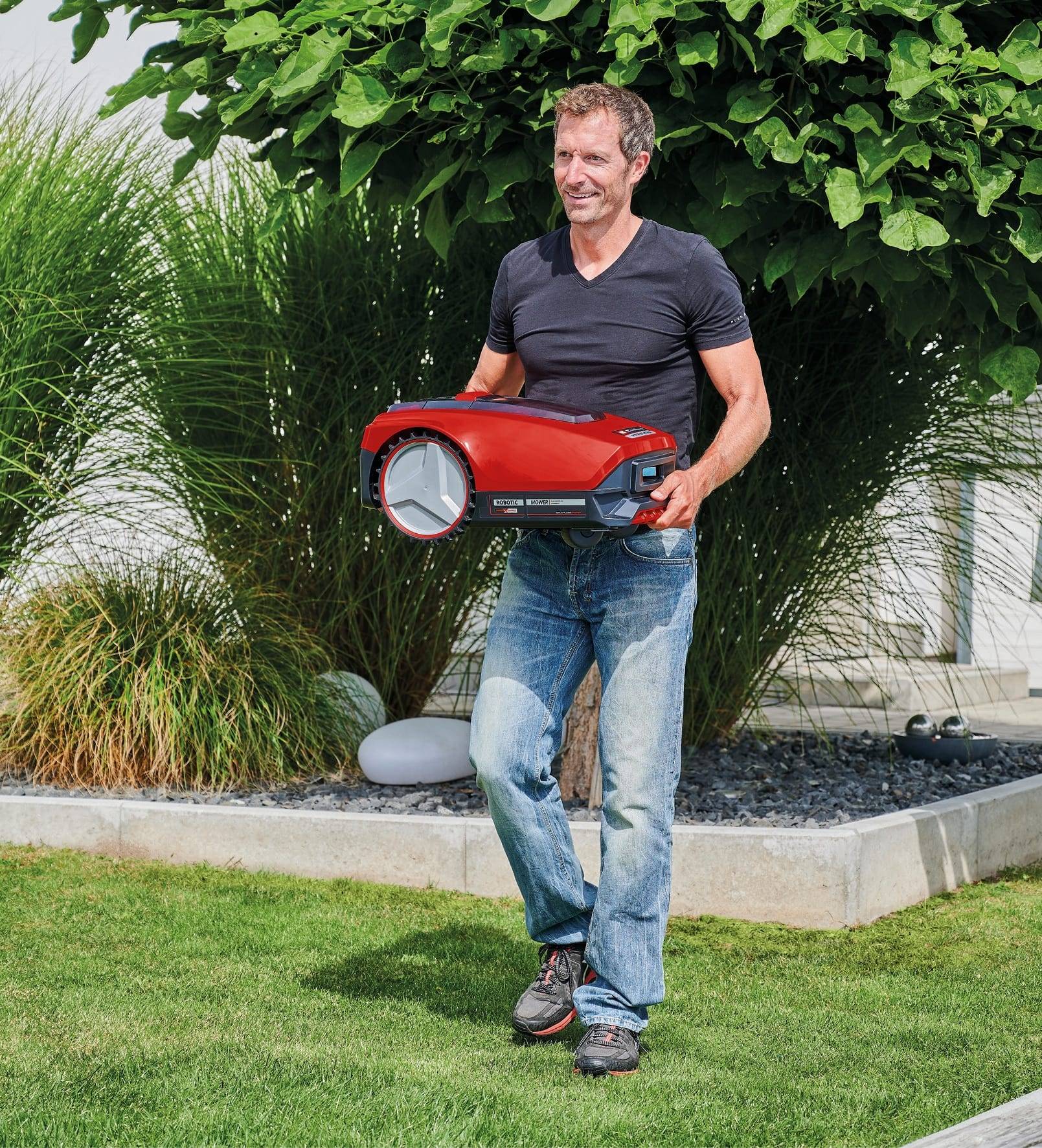 Eine Person geht auf Gras und hält einen roten robotischen Rasenmäher, umgeben von grünen Pflanzen und einem Baum.