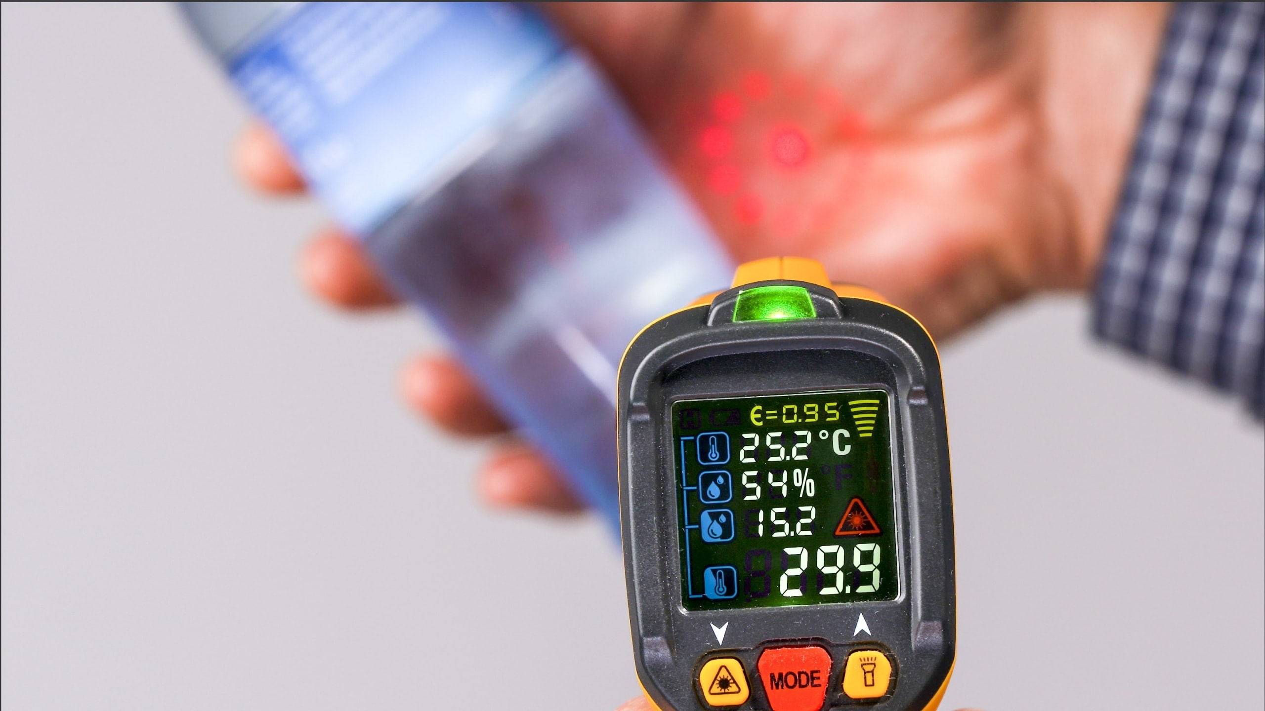 Ein tragbares Infrarot-Thermometer zeigt eine Temperatur von 29,9°C auf einem Bildschirm an, mit unscharfem Hintergrund und rotem Laser-Punkt auf einer Hand.