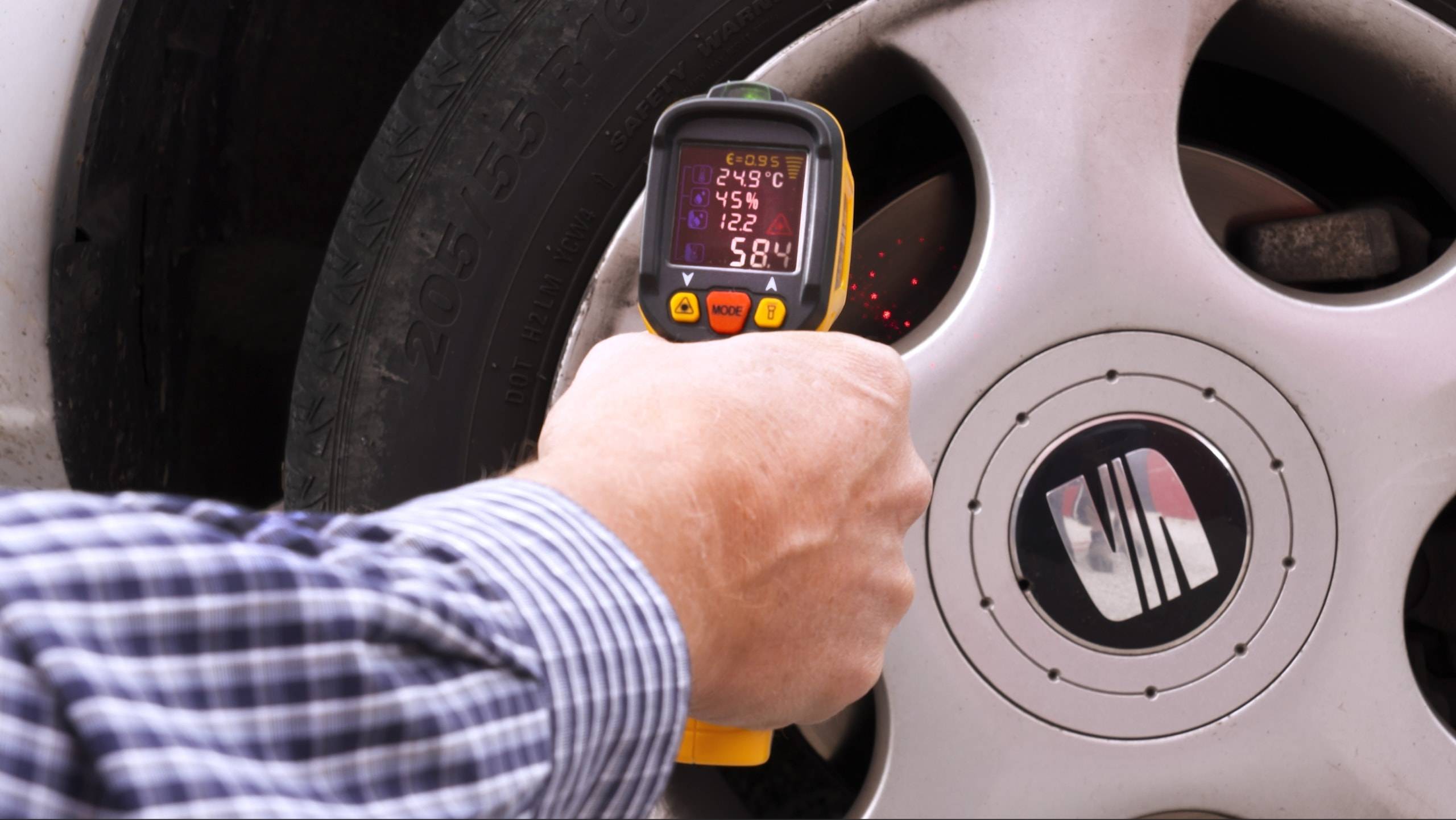 Eine Person hält ein digitales Thermometer neben einen Autoreifen und zeigt Messungen an: '24,9°C', '142', '5,8'. Die Felge des Reifens ist sichtbar.