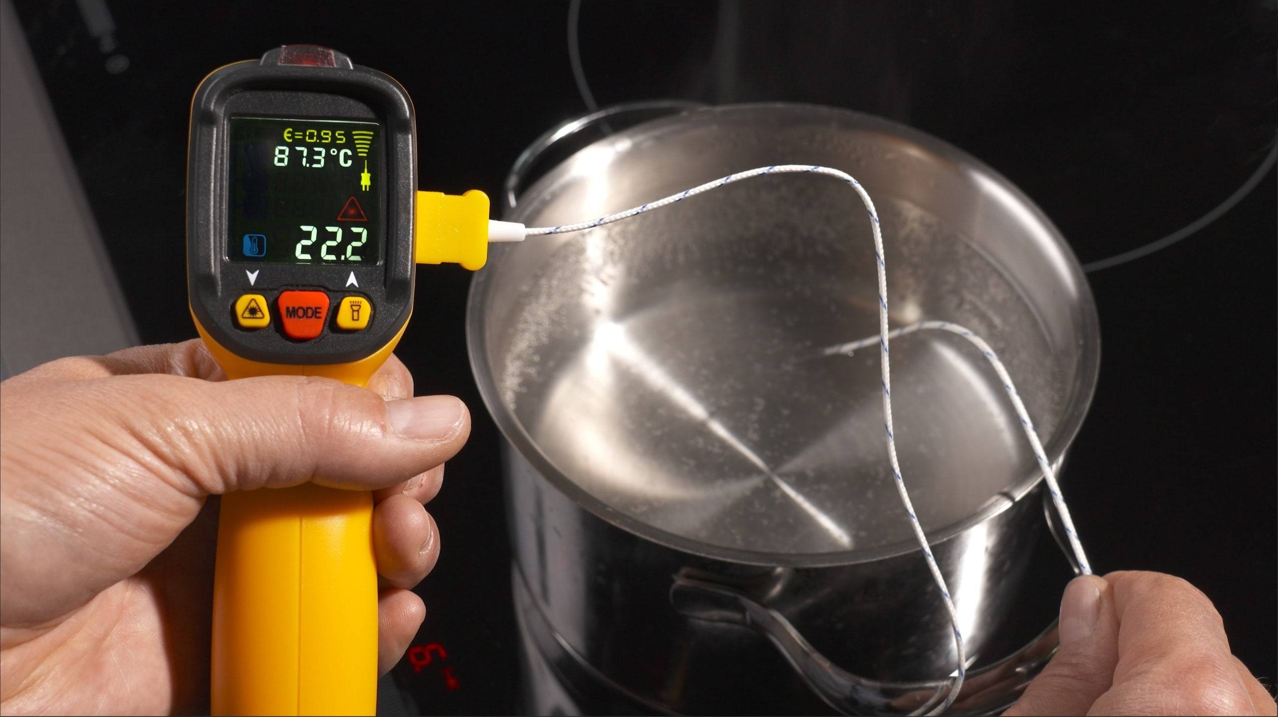 Eine Hand hält ein Thermometer, das 22,2°C anzeigt, über einem Topf mit kochendem Wasser und zeigt damit einen Temperaturmessvorgang an.