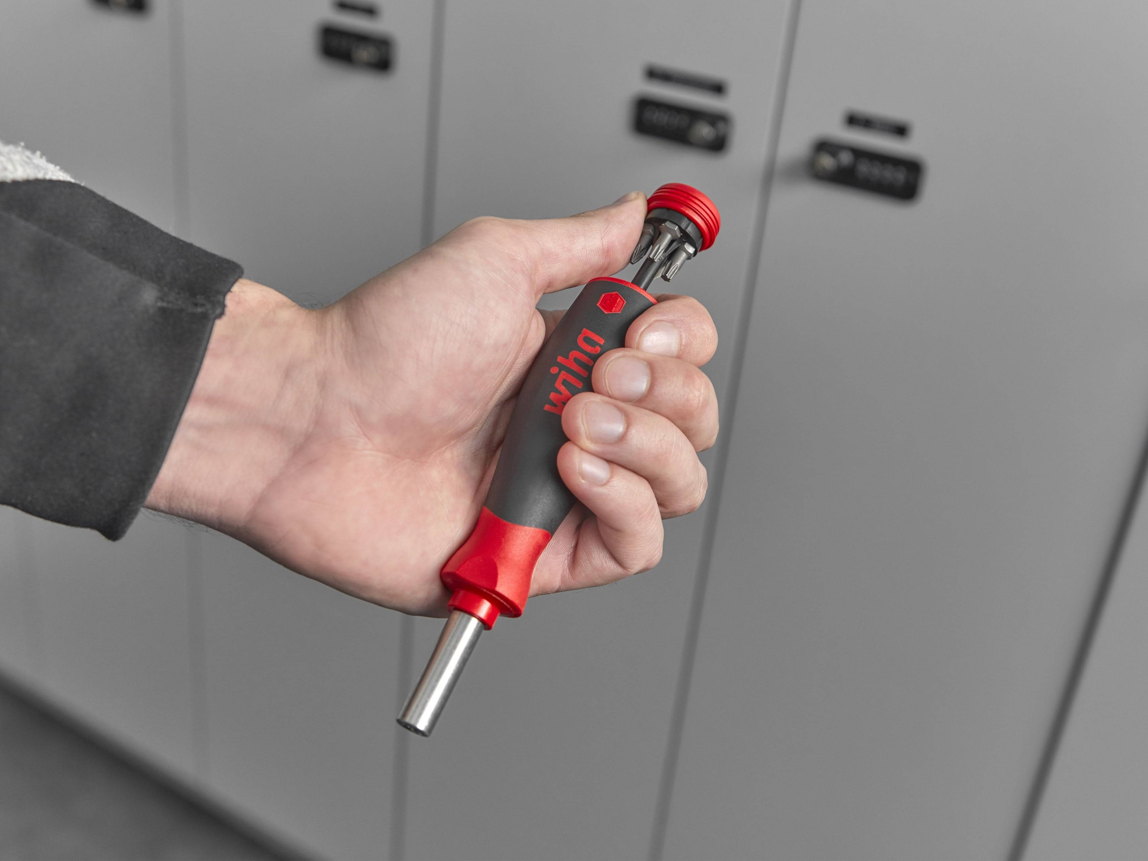 Eine Hand hält einen Schraubenzieher mit einem rot-grauen Griff vor einer Reihe von Spinden.