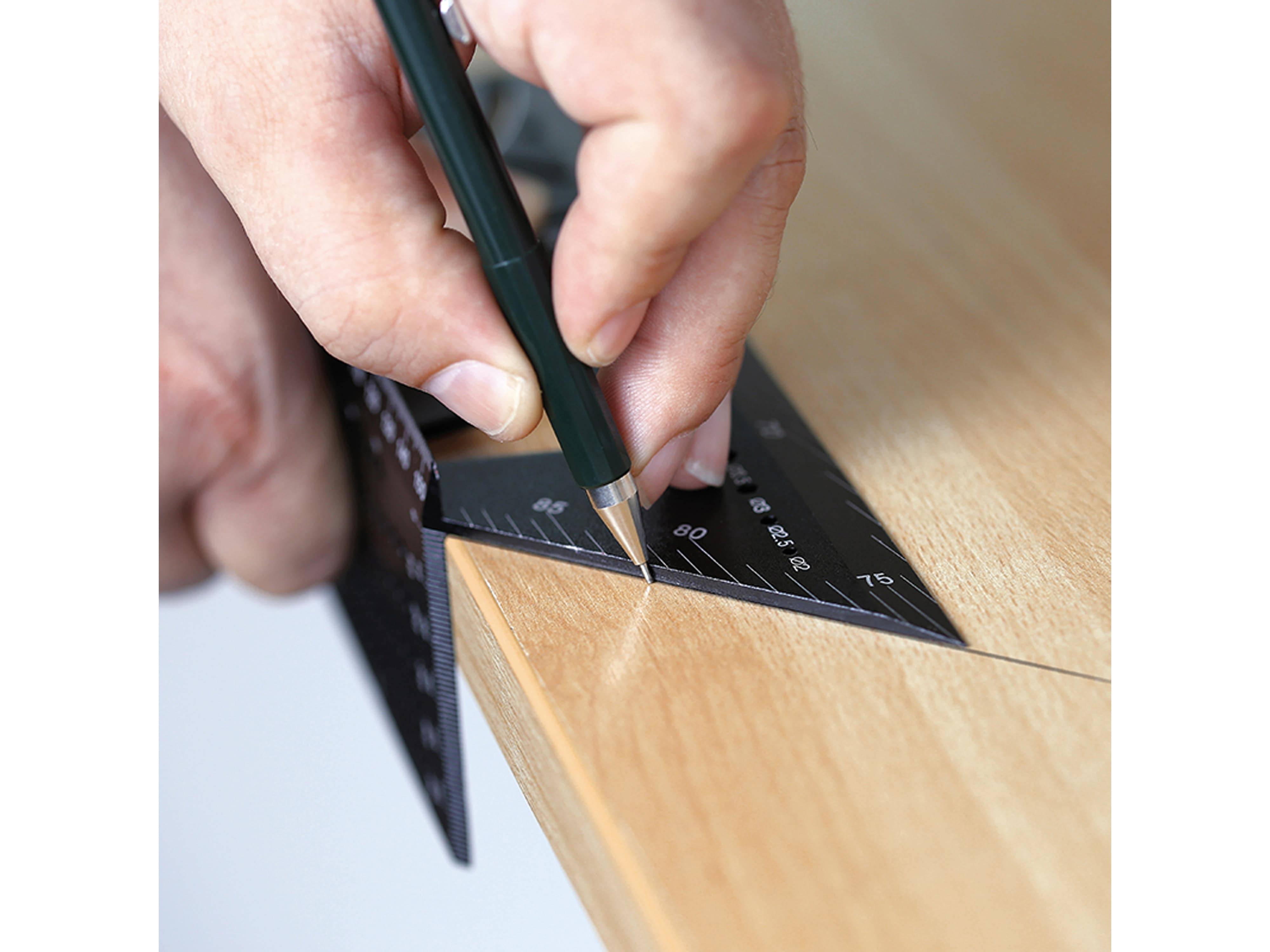 Eine Hand verwendet ein schwarzes dreieckiges Lineal und einen grünen Stift, um eine präzise Linie auf einer Holzoberfläche zu ziehen, was Messung und Planung anzeigt.