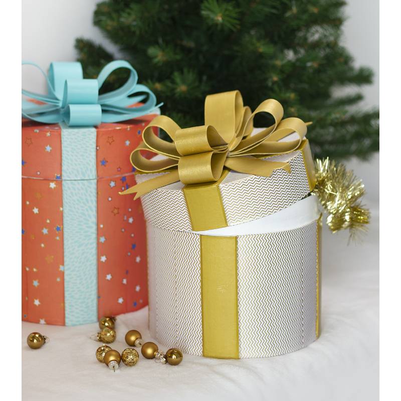 Zwei eingepackte Geschenkboxen mit Schleifen vor einem Weihnachtsbaum; eine ist gold und weiß, die andere rot mit Sternen.