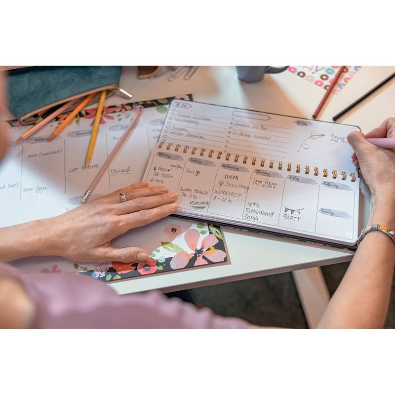 Eine Person schreibt in einen kleinen Spiralplaner auf einem Tisch mit bunten Buntstiften und Blumenpapieren und plant die Woche mit Aufgaben und Notizen.