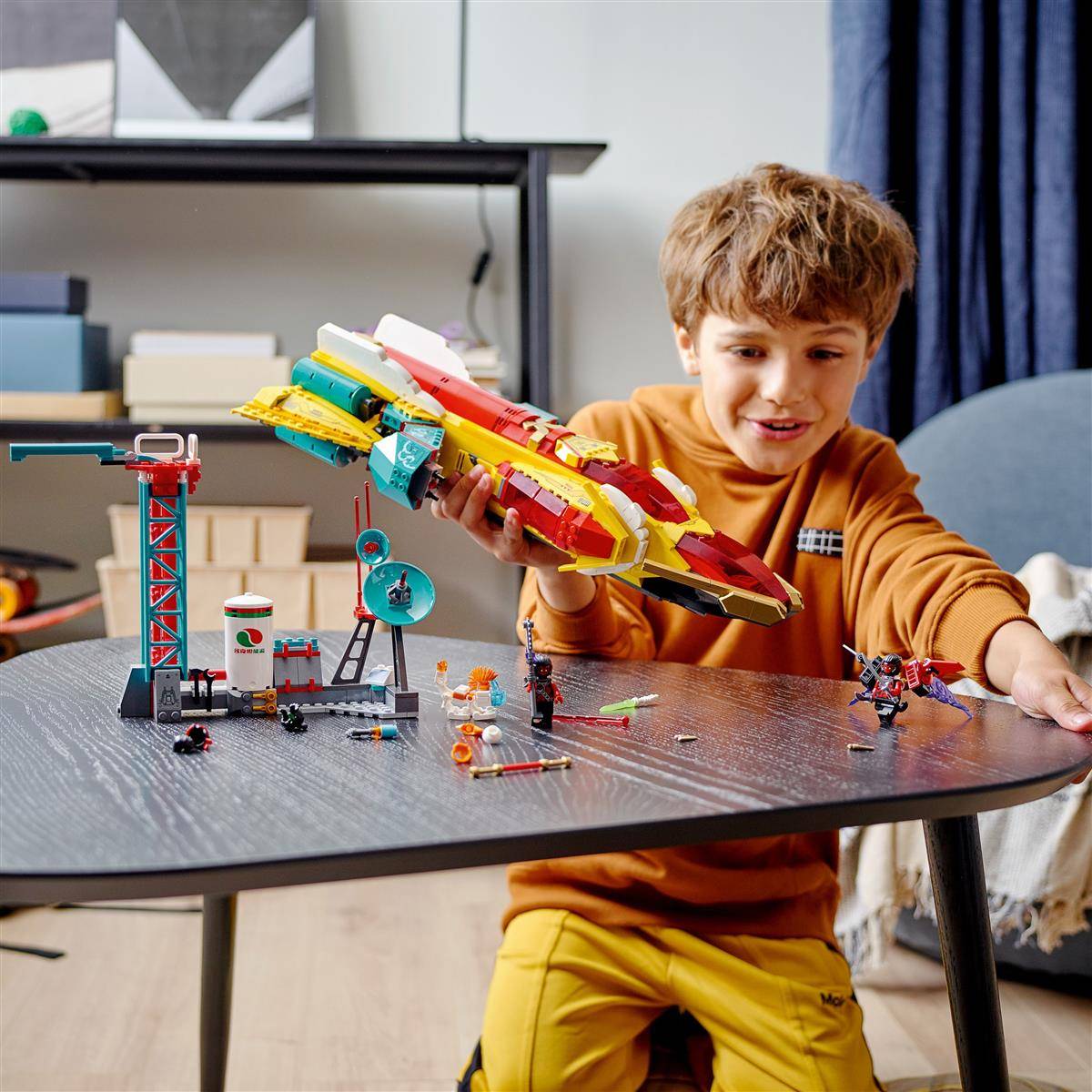 Ein Kind spielt fröhlich mit einem bunten Spielzeug-Raumschiff auf einem Tisch, umgeben von verschiedenen kleinen Spielzeugfiguren und Strukturen.