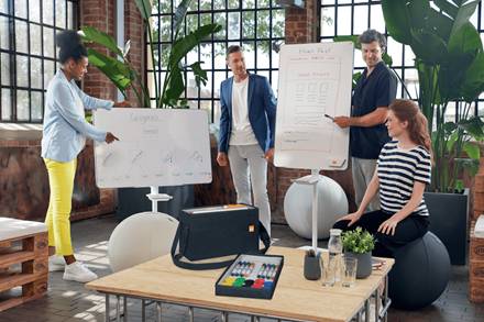 # Team präsentiert Geschäftsideen in einem hellen Büro, mit zwei Personen an Whiteboards und einem sitzenden Kollegen, der während eines Meetings zusieht.