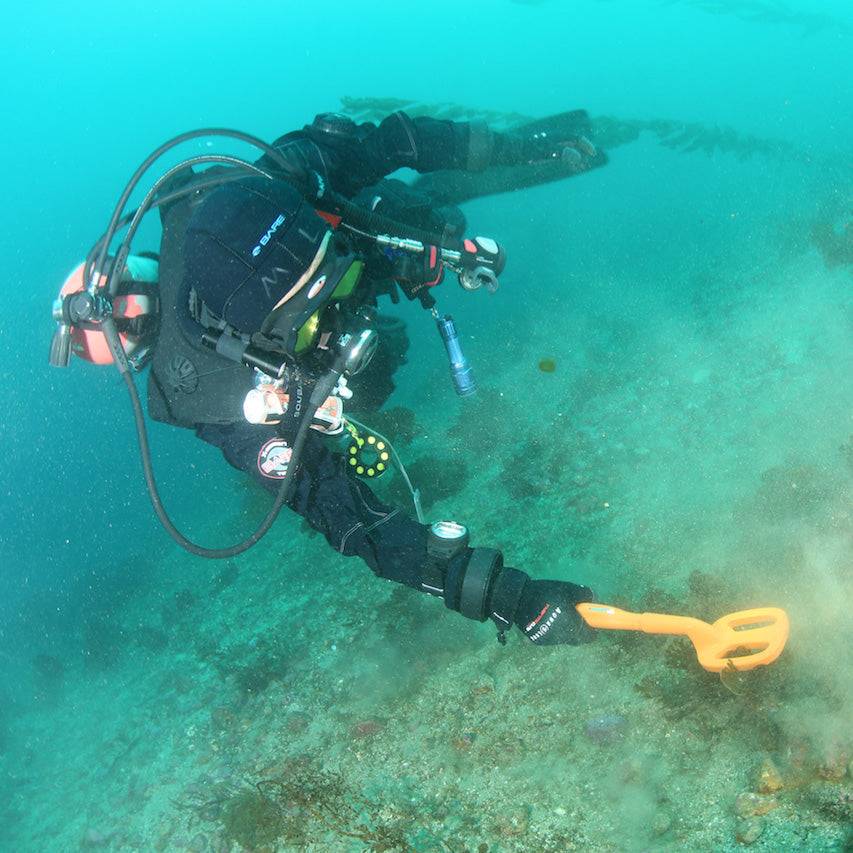 Taucherin unter Wasser, die den Meeresboden mit einem tragbaren Metalldetektor und einem orangefarbenen Grabwerkzeug abtastet.