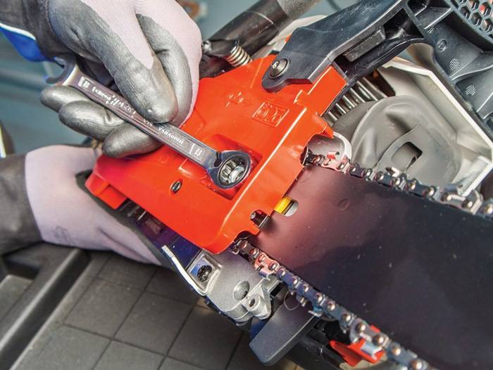 Eine Handschuh tragende Hand benutzt einen Schraubenschlüssel, um die Spannung am roten Sägekettenlaufrad einzustellen. Der Fokus liegt auf den Details des Werkzeugs und der Maschine.