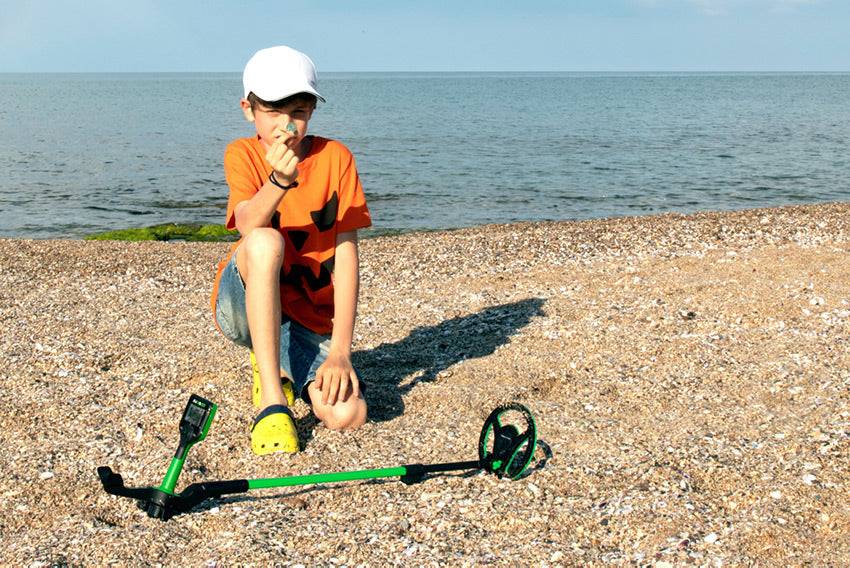 Junge, der auf einem Kieselstrand neben einem Metalldetektor kniet und einen kleinen Fund in der Nähe des Meeres hält.