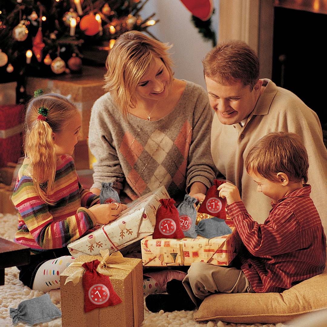 Familie von vier Personen öffnet Weihnachtsgeschenke auf dem Boden neben einem geschmückten Baum und einem Kamin.