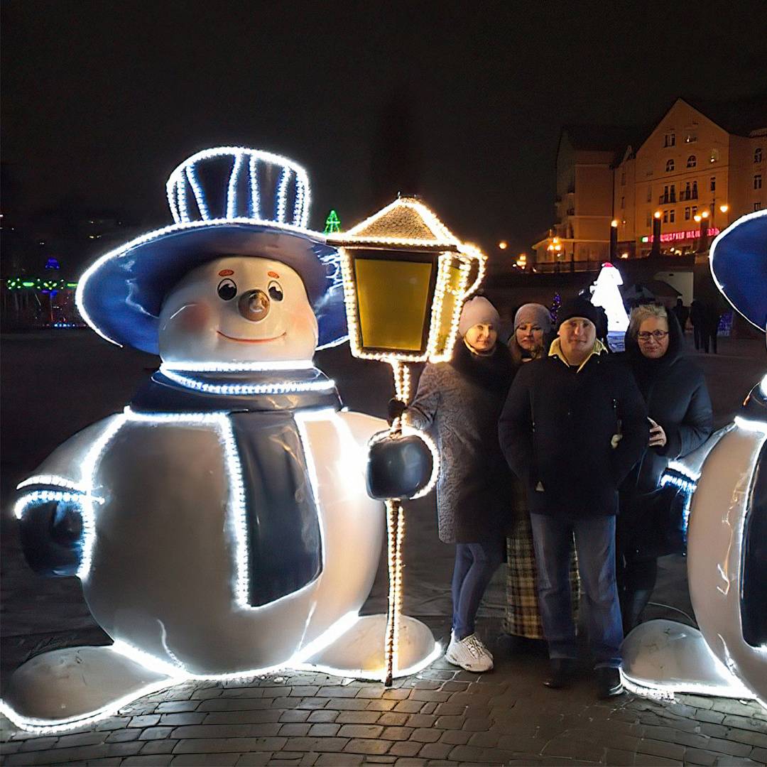 Vier Personen posieren neben einer beleuchteten Schneemann-Weihnachtsdekoration, die eine Laterne hält, auf einem nächtlichen Stadtplatz.