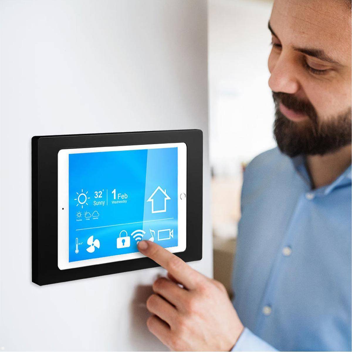 Eine Person interagiert mit einem Smart Home Bedienfeld, das an der Wand montiert ist. Der Bildschirm zeigt Wetterinformationen an: „32°, Sonnig, 1. Feb, Mittwoch.