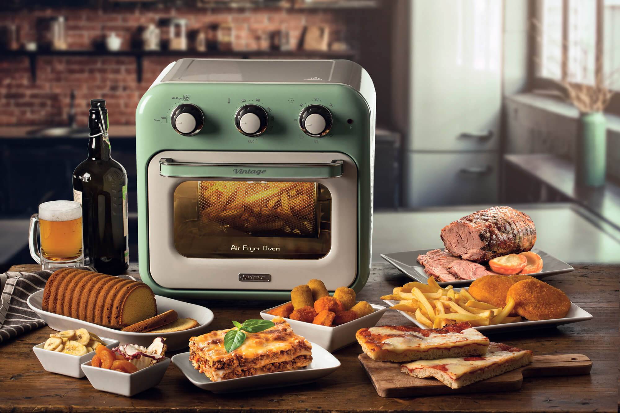 Retro-grüner Heißluftfritteuse-Ofen auf einem Küchentisch umgeben von gekochtem Essen, Brot, Sandwiches, Pommes Frites, Gebäck, Bierflasche und einem Becher Bier.