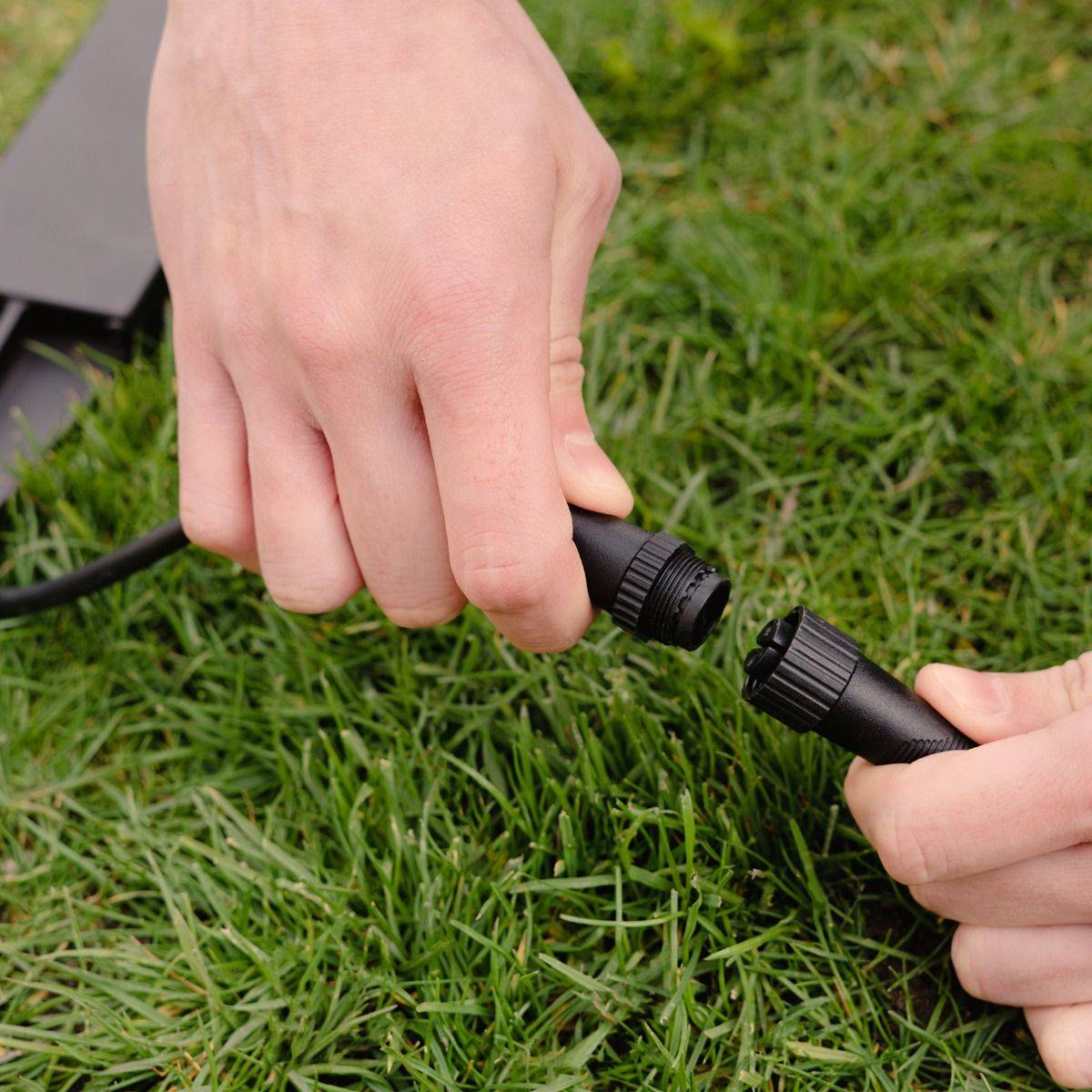 Hände verbinden zwei schwarze Kabelstecker über Gras und verbinden wahrscheinlich ein Outdoor-Stromkabel oder ein Gartenstromkabel.