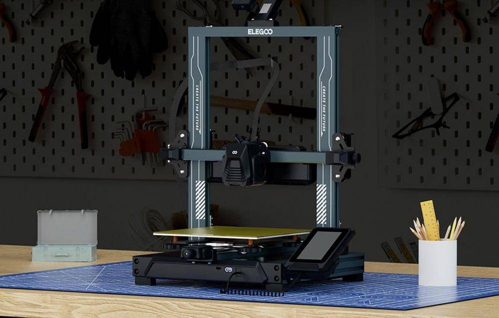 Ein 3D-Drucker steht auf einem Tisch und ist einsatzbereit. Er verfügt über ein Dual-Rail-Design und eine Touchscreen-Schnittstelle. Eine Werkstattumgebung ist im Hintergrund zu sehen.