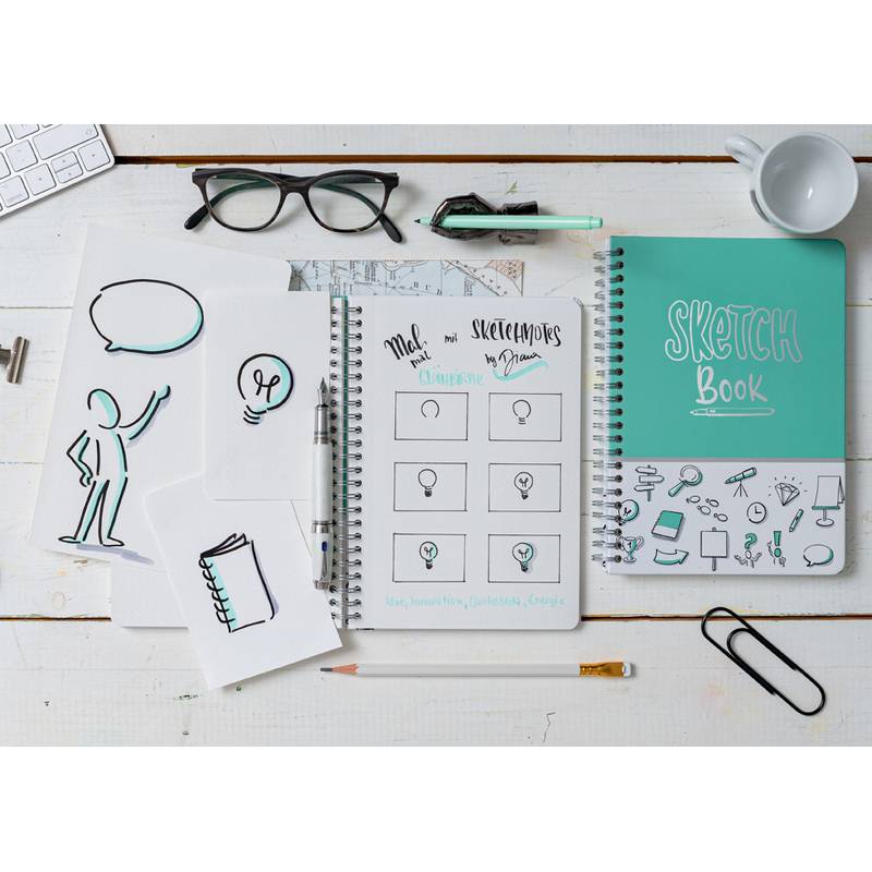 Ein Arbeitsbereich mit einem Skizzenbuch, auf dem sechs Glühbirnenzeichnungen, eine Figurenskizze, eine Brille, eine Tastatur und verschiedene Schreibwaren zu sehen sind.