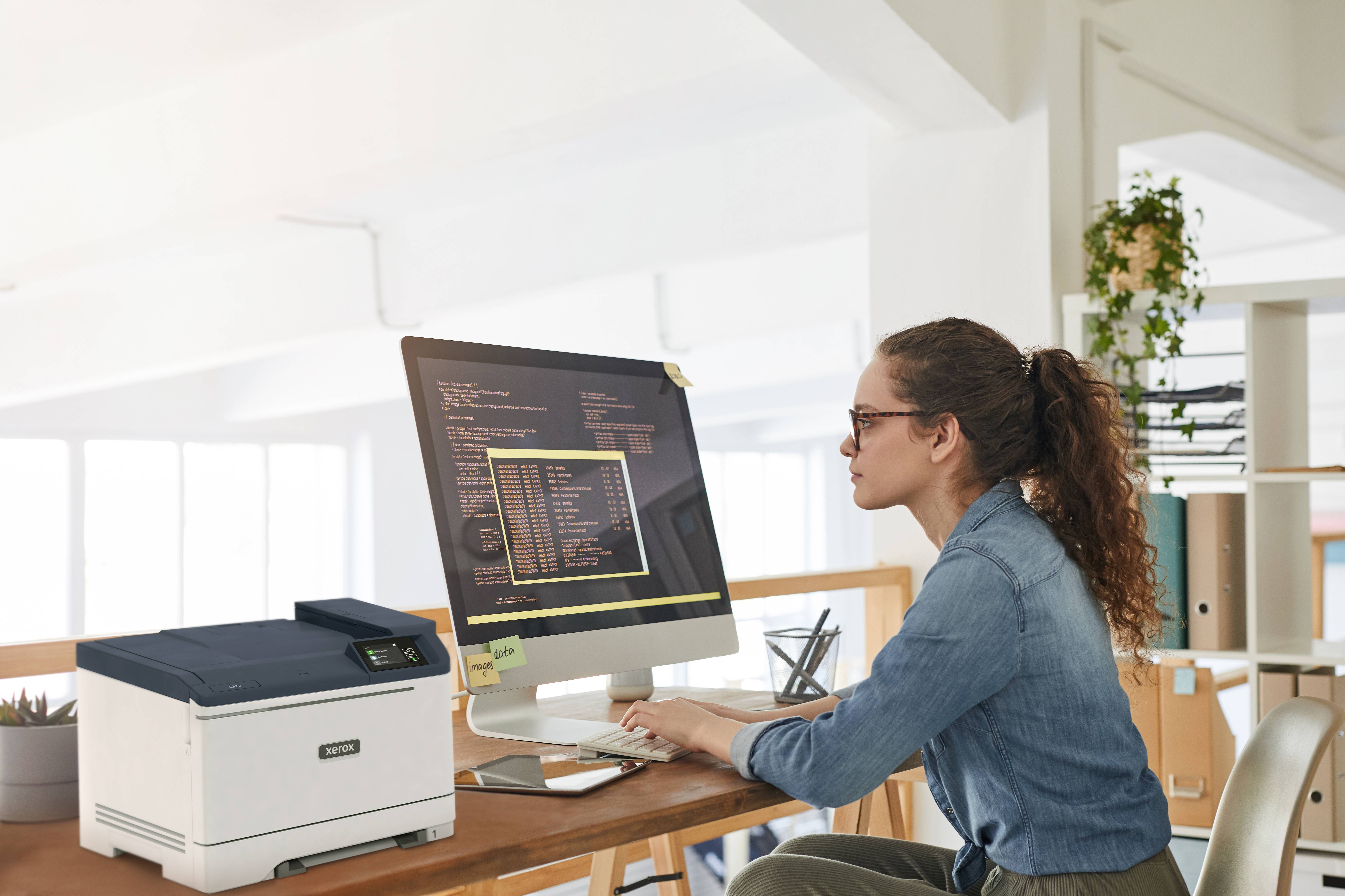 Frau am Schreibtisch, die auf einem Desktop-Computer neben einem Drucker in einem hellen Büroarbeitsraum tippt.