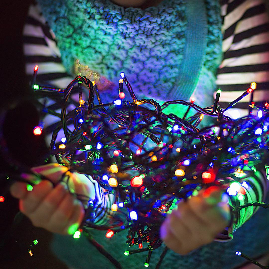 Person, die ein verworrenes Bündel aus farbenfrohen Weihnachtslichterketten mit vielen leuchtenden Glühbirnen in beiden Händen hält.