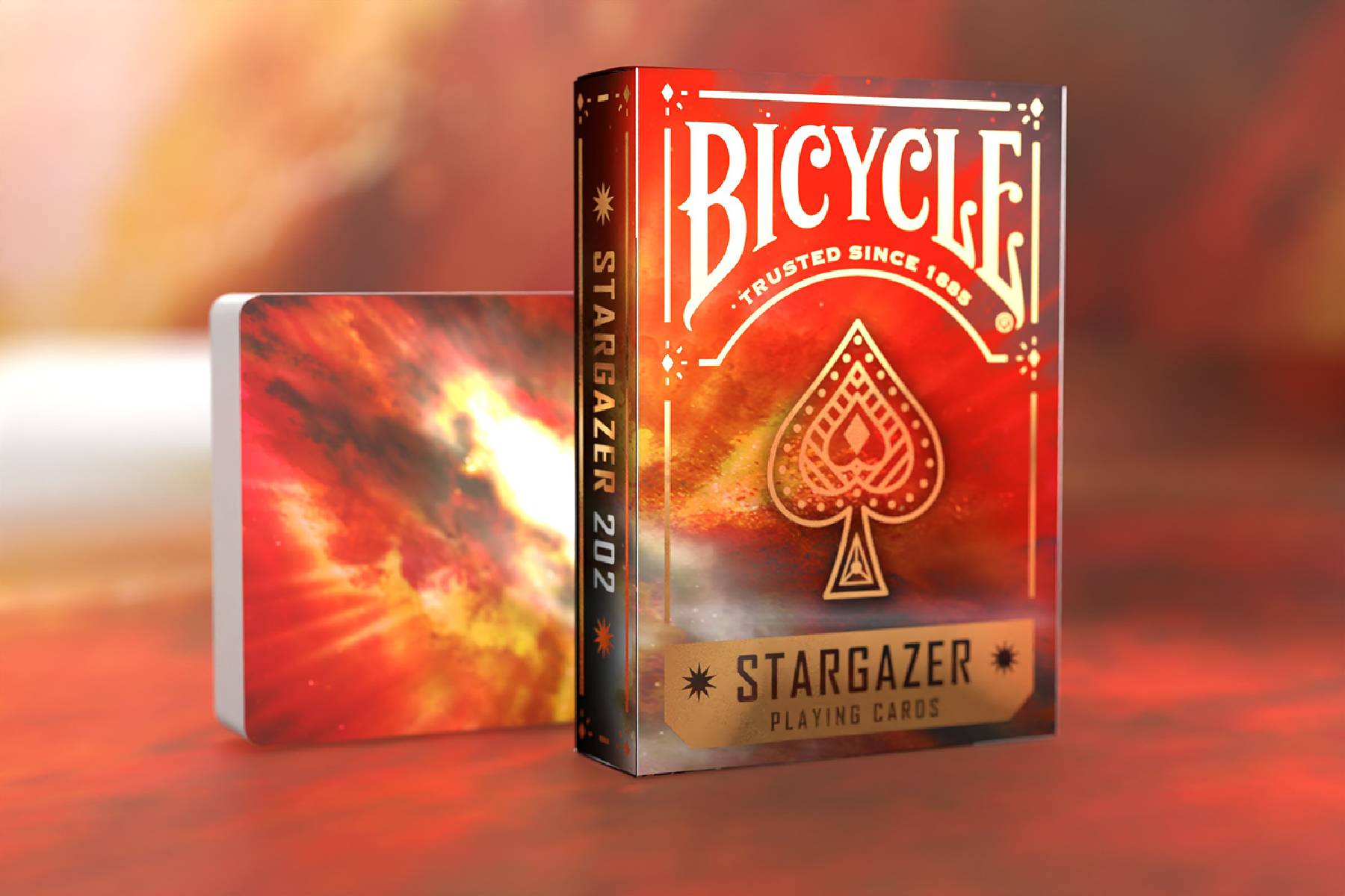 Bicycle Stargazer Spielkartenschachtel mit rot-oranger himmlischer Kunstwerk und passendem Deckschachtel auf verschwommenem warmem Hintergrund.