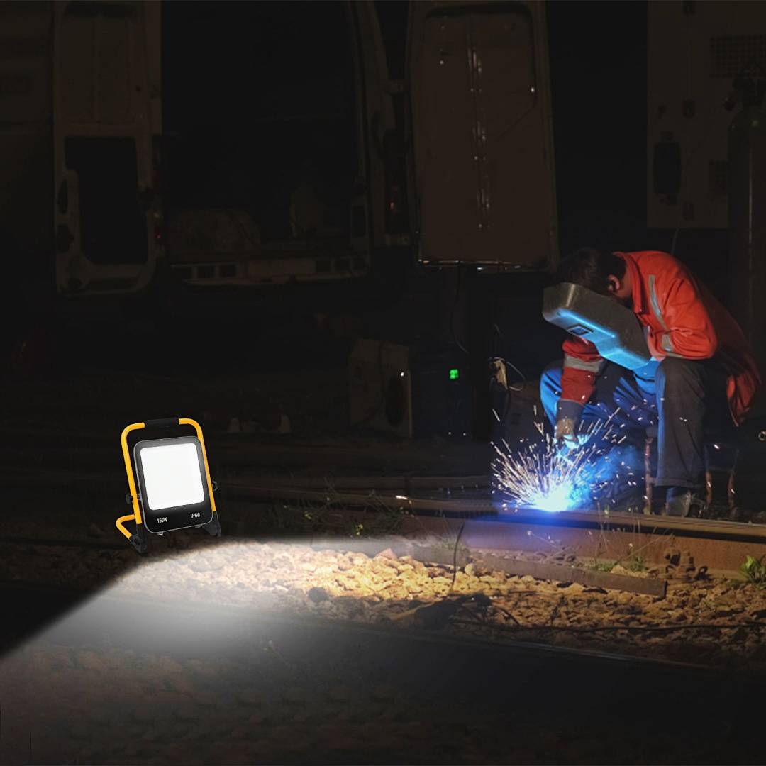 Arbeiter in orangefarbener Schutzausrüstung schweißt nachts an Eisenbahnschienen, während ein tragbares Arbeitslicht die Schienen und das Schotterbett in der Nähe beleuchtet.
