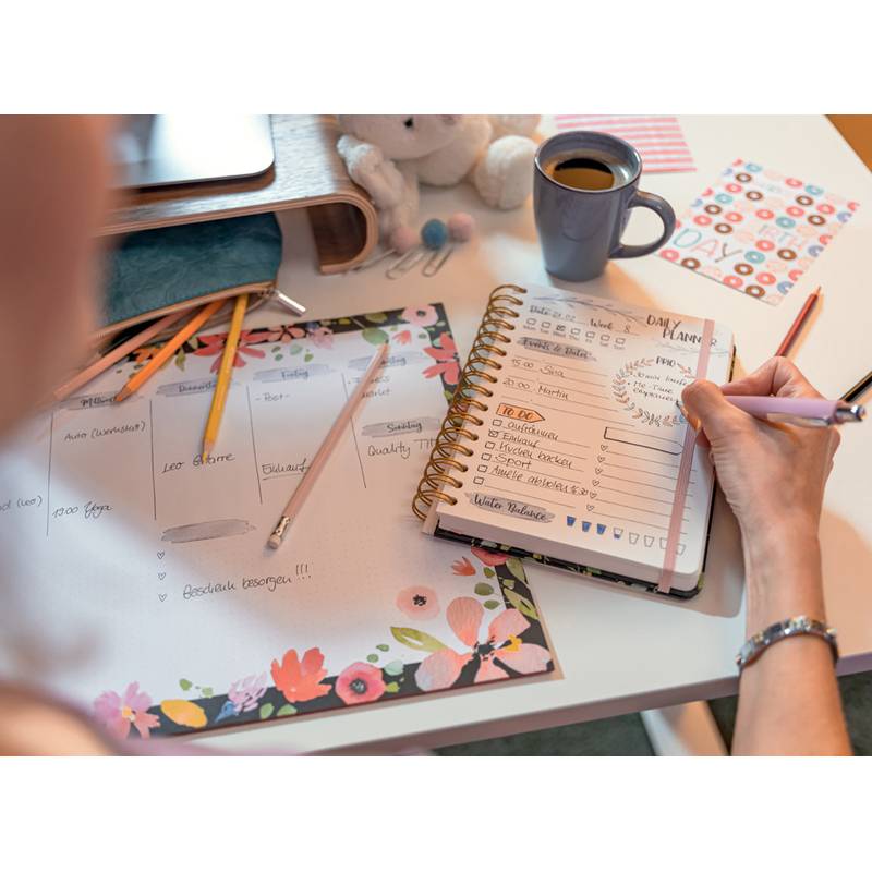 Eine Person schreibt in einem Spiralplaner auf einem Schreibtisch mit Briefpapier mit Blumenmuster, bunten Farbstiften und einer Tasse Kaffee, was auf eine Planungsphase hindeutet.