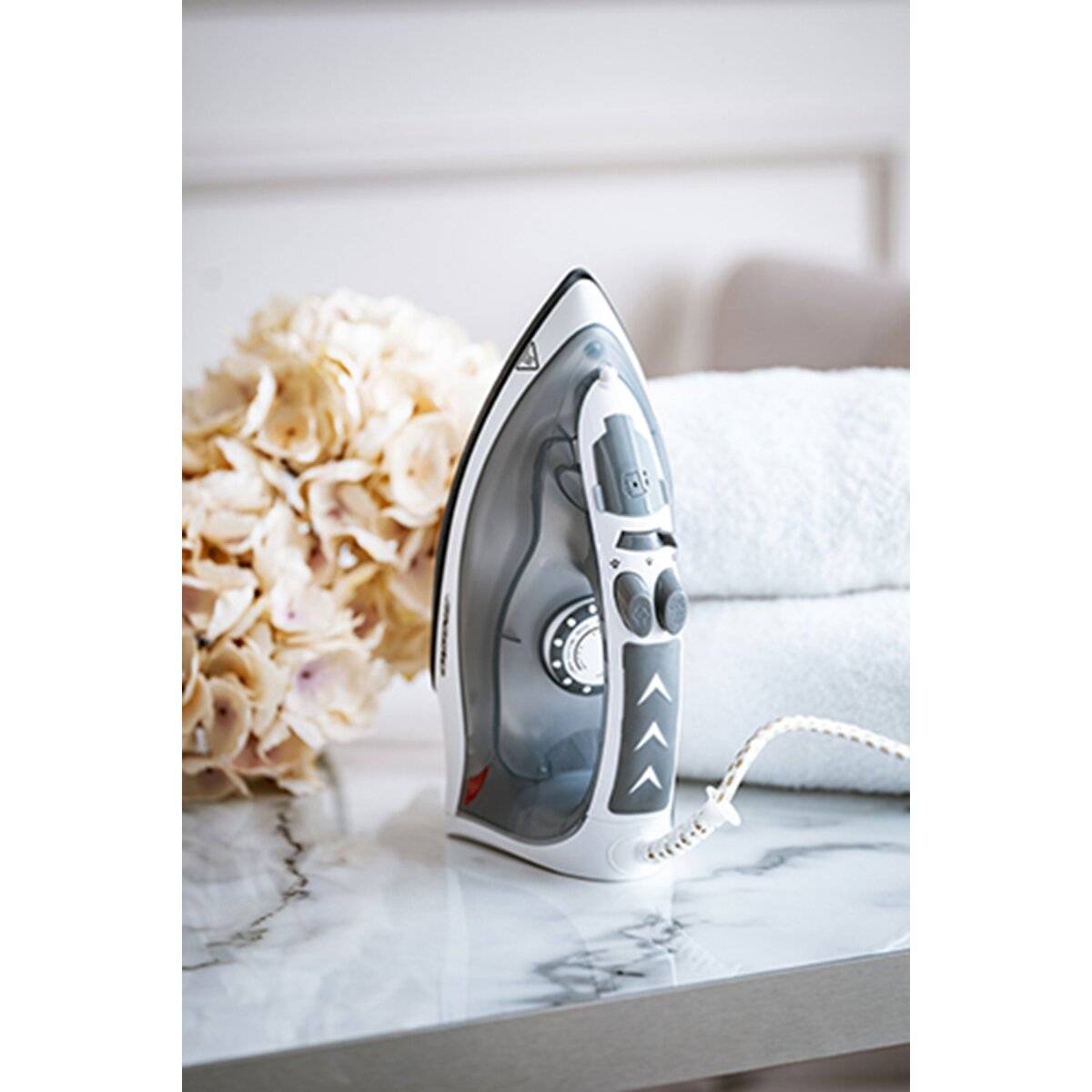 Ein Dampfbügeleisen steht aufrecht auf einer Marmorflache neben gefalteten Handtuchern und einem Strauß hellfarbener Hortensien, was auf Haushaltssor­ge hindeutet.