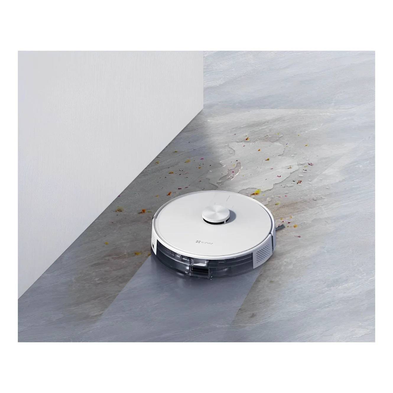 Roboter-Staubsauger reinigt einen grauen Boden neben einer Wand, mit verstreutem Schutt und einer Nassstelle voraus.