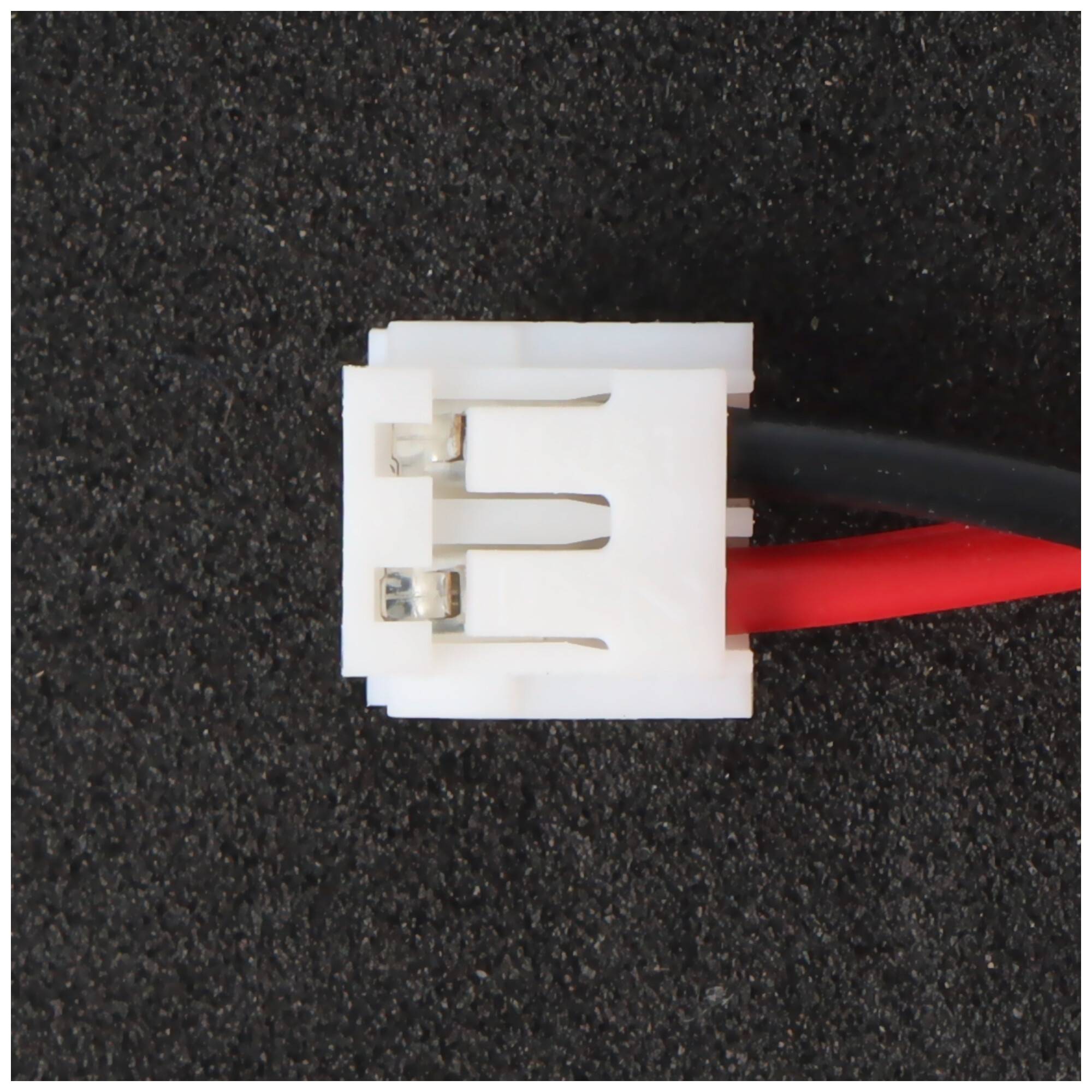 Großaufnahme eines weißen zweipoligen Elektrosteckers mit roten und schwarzen Kabeln auf einer dunklen strukturierten Oberfläche.