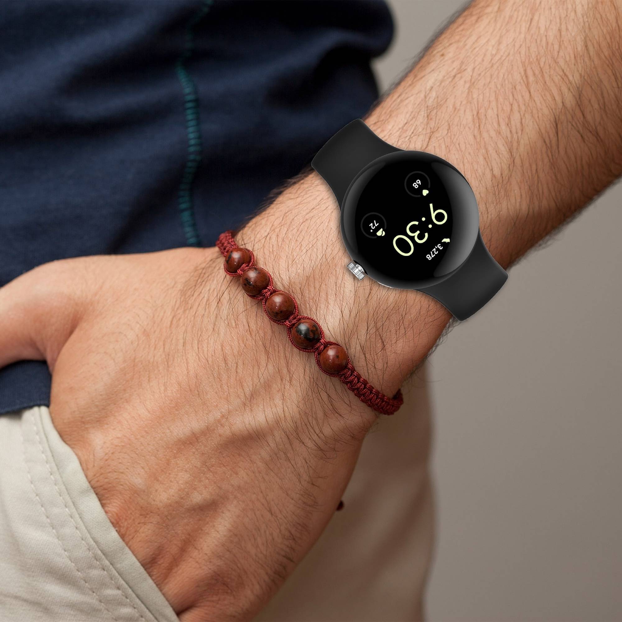 Nahaufnahme eines Handgelenks, das eine schwarze runde Smartwatch mit digitaler Zeitanzeige und ein rotes Perlenarmband trägt.
