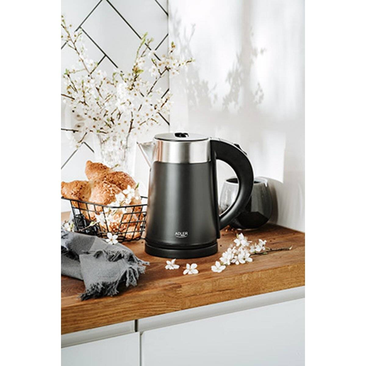 Ein schwarzer elektrischer Wasserkocher steht auf einer Holzarbeitsplatte neben einem Korb mit Croissants und weißen Blumen, mit einer weiß gefliesten Rückwand.