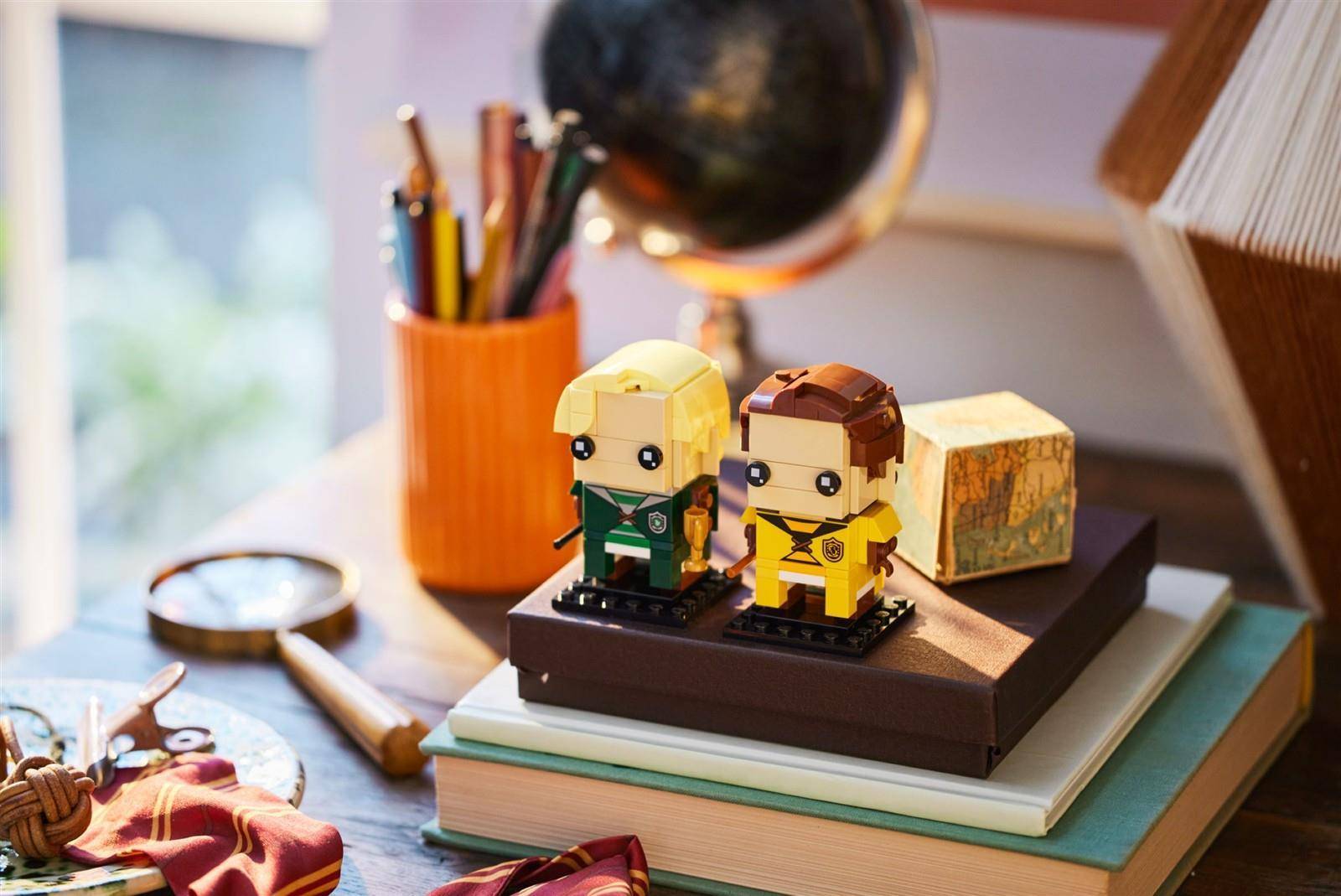 Zwei kleine LEGO-ähnliche Figuren stehen auf gestapelten Büchern neben einem Stiftbecher, einer Lupe und einem Globus auf einem Schreibtisch.